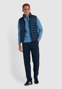 Marineblue gewatteerde vest over een lichtblauwe sweatshirt, gecombineerd met donkere broek en zwarte sneakers. Heeft een ritsluiting en ribgebreide manchetten.