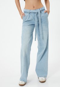 Jeans de tiro alto y pierna ancha en azul claro, con cinturón y bolsillos delanteros. Hechos de mezclilla con una textura suave y un acabado desgastado.