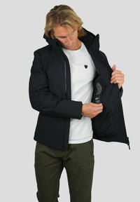 Veste noire isolée avec fermeture éclair et capuche réglable, présentant une poche intérieure. Portée sur un t-shirt blanc et un pantalon olive.
