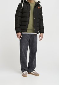 Mann in schwarzer Steppjacke, olivgrünem Sweatshirt, weiten dunklen Jeans und grauen Sneakers steht vor weißem Hintergrund.