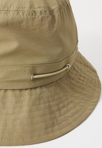 Beige bøttehat lavet af letvægtsstof, med en bred, flad skygge og en syet detalje. Inkluderer en dekorativ løkke på siden.