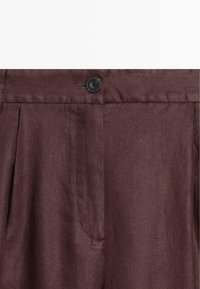 Pantalon marron foncé taille haute avec fermeture par bouton noir et plis à l'avant, montrant la texture du tissu et les détails des coutures.