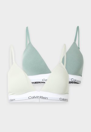 Žena stojaca bosá, oblečená v bielej podprsenke Calvin Klein a čiernom spodnom prádle Calvin Klein na jednoliatom svetlom pozadí.