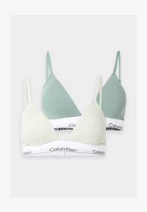 Kobieta stojąca boso, ubrana w biały biustonosz Calvin Klein i czarną bieliznę Calvin Klein na jednolitym jasnym tle.