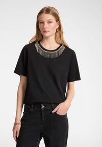 Camiseta de manga corta negra con cuello redondo, que presenta un diseño decorativo de flecos plateados en el cuello. Textura de tela suave.