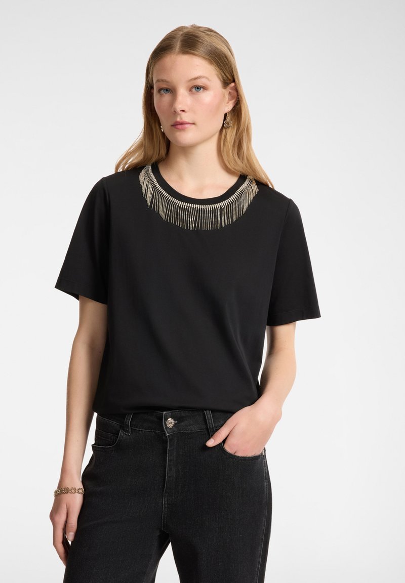 Camiseta de manga corta negra con cuello redondo, que presenta un diseño decorativo de flecos plateados en el cuello. Textura de tela suave.