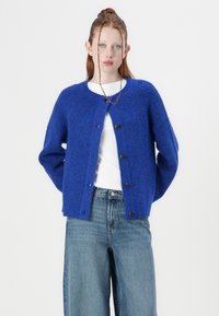 Blue knitted cardigan with a round neckline, long sleeves, and five large buttons. Textured fabric with a soft appearance. Paired with blue jeans.