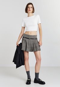 Giovane donna con top corto bianco, gonna mini a pieghe a quadri con cintura nera, calze grigie e mocassini neri, che tiene un giubbotto nero.