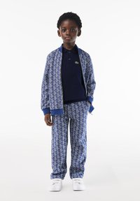 Niebieski dres z geometrycznymi wzorami, składający się z zapinanej na zamek kurtki oraz dopasowanych spodni. Białe sneakersy uzupełniają całość.