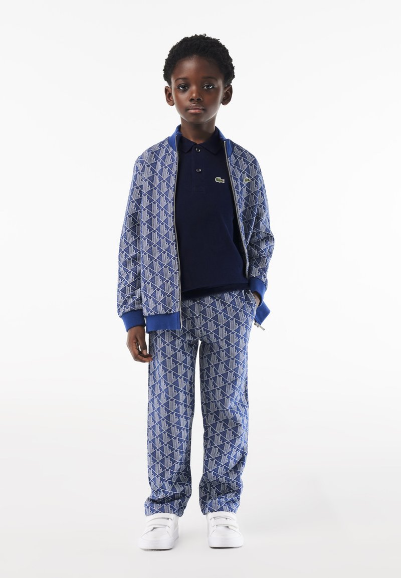 Niebieski dres z geometrycznymi wzorami, składający się z zapinanej na zamek kurtki oraz dopasowanych spodni. Białe sneakersy uzupełniają całość.