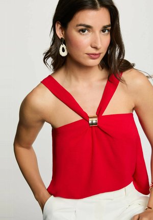 Donna con capelli scuri che indossa una canotta rossa con fibbia dorata e pantaloni bianchi, posa con una mano in tasca su sfondo grigio.