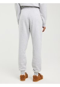 Asmuo, dėvintis šviesiai pilkas sportines kelnes ir rudus sportinius batelius, stovintis atsisukęs nugarą priešais paprastą baltą foną.