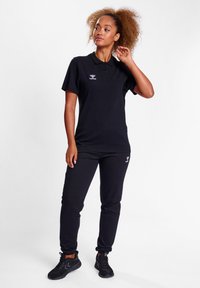 Czarna koszulka polo z krótkim rękawem i kołnierzykiem, w zestawie z czarnymi spodniami dresowymi, które mają logo na klatce piersiowej i biodrze. Czarne buty sportowe.