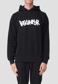 Persona che indossa un hoodie nero con il testo bianco in stile graffiti "DISCLAIMER" sul petto, abbinato a pantaloni neri su uno sfondo neutro.