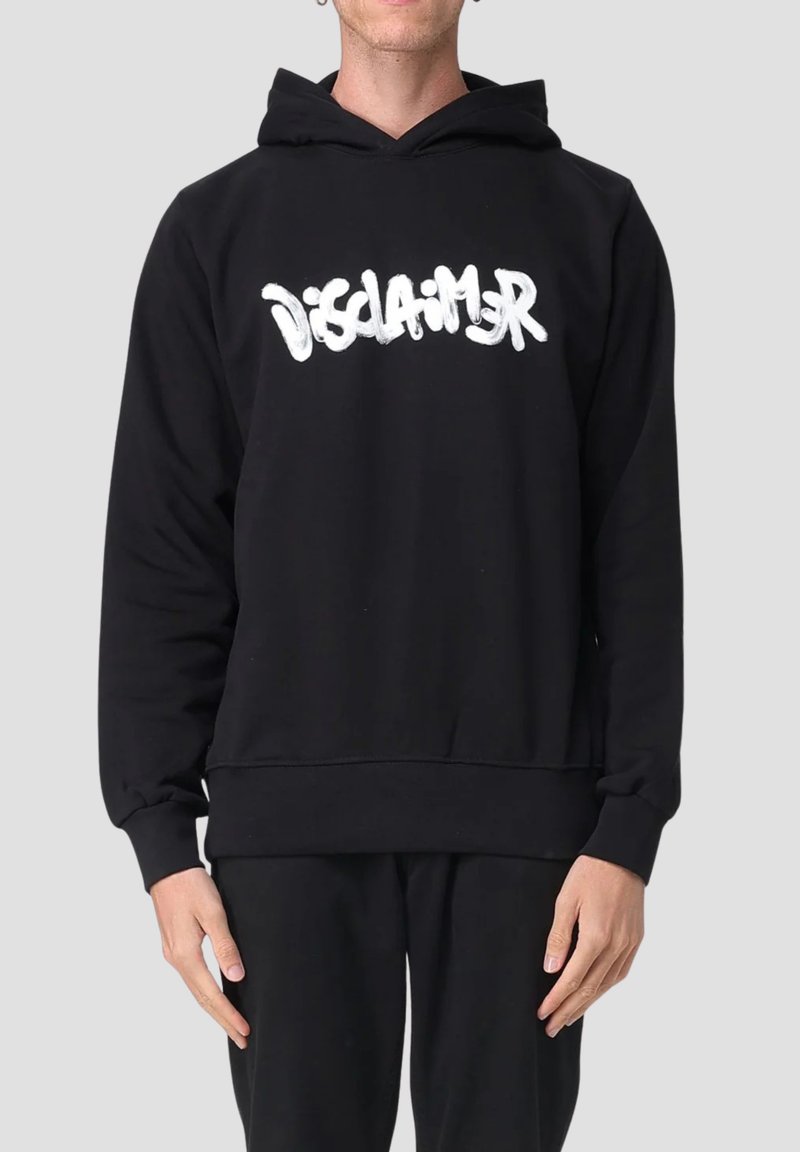 Persona che indossa un hoodie nero con il testo bianco in stile graffiti "DISCLAIMER" sul petto, abbinato a pantaloni neri su uno sfondo neutro.