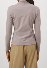 Långärmad topp i ljus lavendelfärgad tyg med mockkrage, slät textur och figursydd design, i kombination med beiga högmidjade byxor.