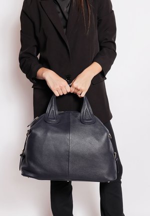 Vrouw in een zwarte colbert en broek houdt een grote marineblauwe handtas van textuurleer vast bij de handvaten op taillehoogte.