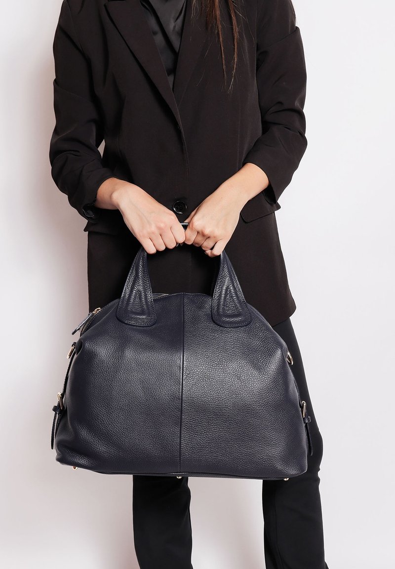 Una donna che indossa un blazer nero e pantaloni tiene una grande borsa in pelle blu navy testurizzata per i manici all'altezza della vita.