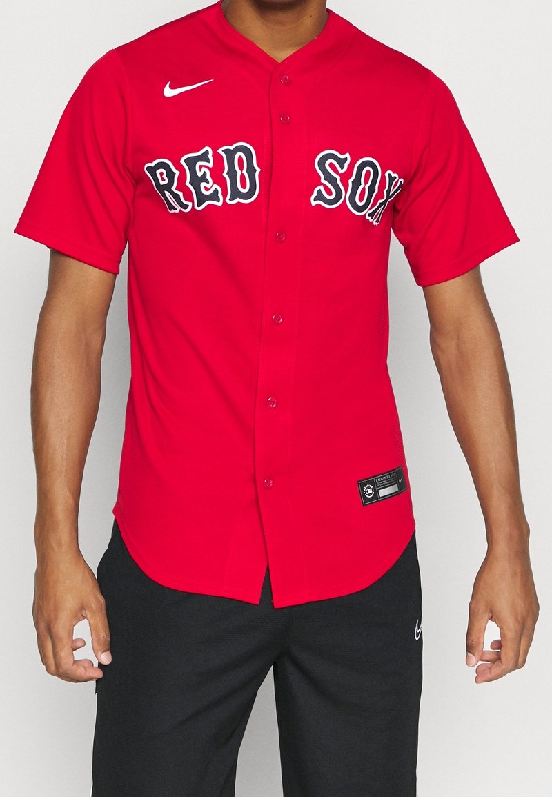 Homme portant un maillot de baseball rouge des Boston Red Sox avec un pantalon noir sur un fond uni.
