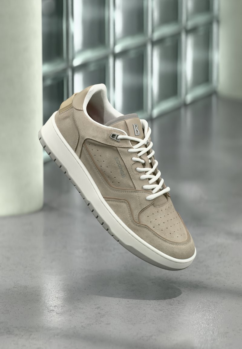 Baskets en daim beige avec lacets et semelle blanche, bout perforé et logo de la marque, suspendues au-dessus d'un sol gris poli près de blocs de verre.