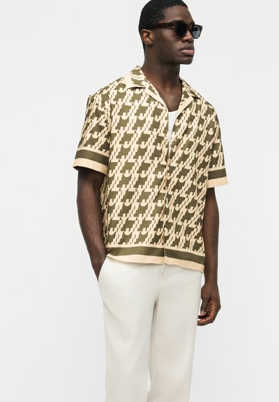 Άνδρας που φοράει καφέ και μπεζ πουκάμισο με κοντά μανίκια και σχέδιο, άσπρο παντελόνι και σκούρα γυαλιά ηλίου, στέκεται με τα χέρια στις τσέπες.