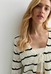 Weißer Strickcardigan mit schwarzen horizontalen Streifen, der ein Spitze-Muster hat, einen V-Ausschnitt und einen gebundenen Frontverschluss über einem passenden Oberteil.