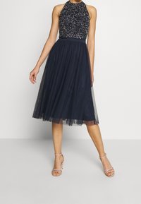 Laivansinis suknelė be rankovių, dekoruota karoliukais viršutine dalimi, su prigludusia liemene ir pūsta, permatomos tiulio sijonu. Papildyta rožinės spalvos aukštakulniais su dirželiais.