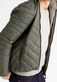 Giacca leggera imbottita color verde oliva con trapuntatura a zig zag, chiusura frontale con cerniera e tasche laterali, realizzata in un materiale sintetico liscio.