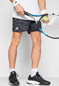 Mustrikud mustad ja hallid spordilühikesed püksid Adidas logo  ja mustade tenniste ja valgete sokkidega, hoides tennisereketit ja pall.