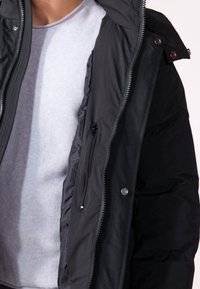 Chaqueta de plumas negra con un bolsillo en el pecho con cremallera, botones de presión y una cremallera robusta, superpuesta sobre un suéter de textura gris claro.