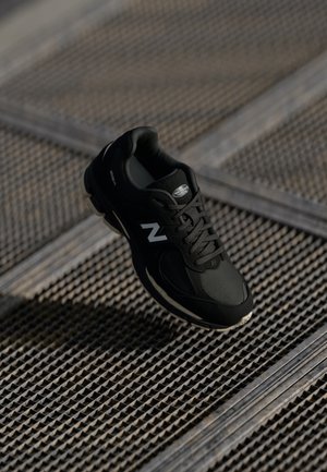 Czarny sneaker New Balance z białym logo i amortyzowaną podeszwą opierający się na fakturowanej metalowej siatce.