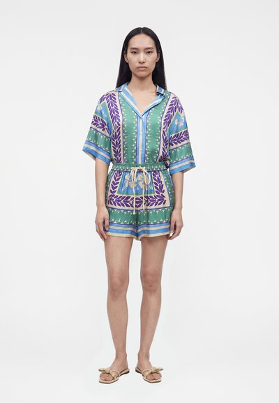 Femme debout face à l'avant, portant une chemise colorée à motifs à manches courtes avec boutons, un short assorti avec taille à cordon et des sandales.