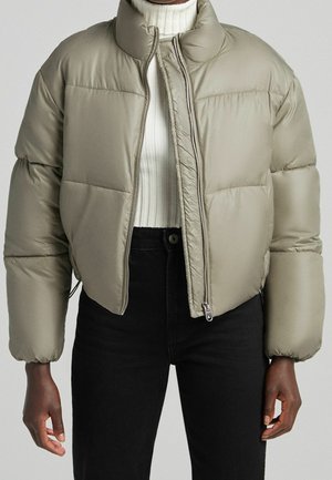 Femme portant une veste courte matelassée beige par-dessus un col roulé côtelé blanc et un pantalon noir taille haute, debout devant un fond uni.