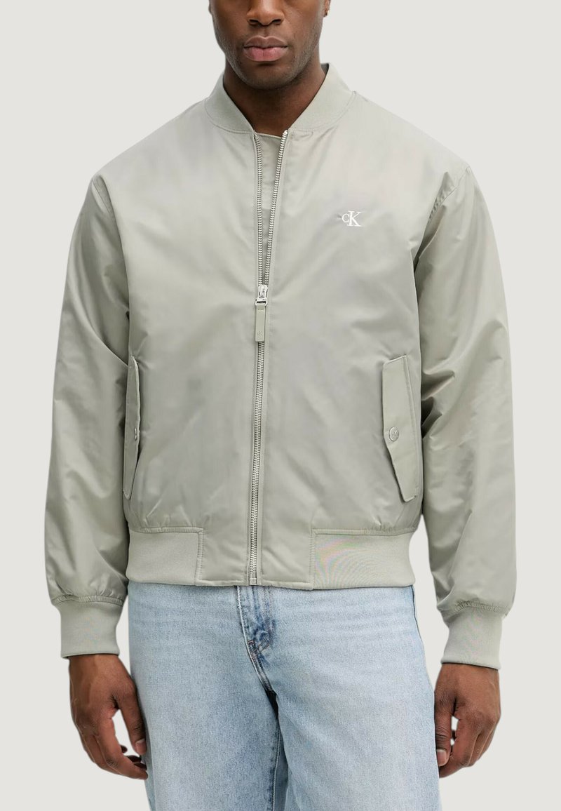 Giacca bomber grigio chiaro con zip frontale, tasche laterali, polsini elasticizzati e un piccolo logo sul petto. Texture liscia, design minimalista.