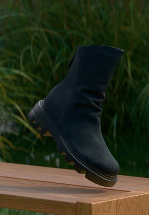 Black mid-calf boot with chunky sole hovers above wooden table, with blurred green foliage in the background.
