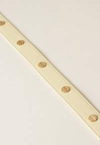Cintura in pelle di colore crema con accenti in metallo dorato a forma circolare distribuiti uniformemente lungo tutta la lunghezza. Texture liscia e linee di design pulite.