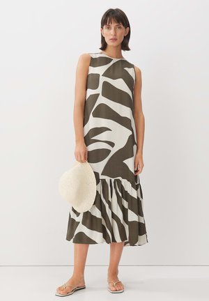 Mouwloze midi-jurk in crème en olijfgroen, met een gelaagde ruchezoom en abstract patroon. Model houdt een geweven strohoed vast.
