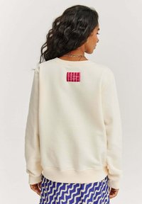 Světlý krémový svetr s ribovaným lemem a manžetami. Na zadní straně se nachází červená a růžová kostkovaná nášivka. Měkká textura, volný střih.