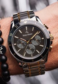 Montre chronographe en métal avec un cadran noir, des accents dorés, des sous-cadrans texturés et un bracelet en acier inoxydable, portée sur un poignet. Bracelet en perles noires à proximité.