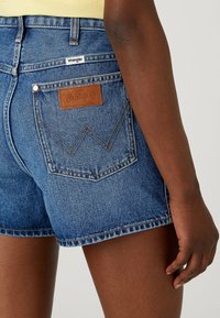 Person som bär blå denimshorts från Wrangler med synlig bakficka och varumärkesetikett, stående mot en enkel ljus bakgrund.