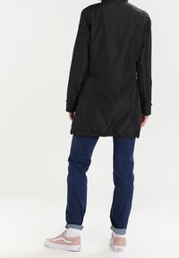 Sort, lang letvægtsfrakke med ståkrave, indsnævret talje og en glat overflade. Båret med blå jeans og lyserøde sneakers.