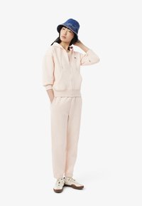 Sudadera rosa clara con cremallera, bolsillos tipo canguro y puños acanalados; combinada con pantalones de chándal rosa claros y zapatillas beige. Sombrero de cubo azul.