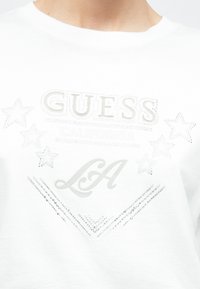 Weißes Hemd mit geprägtem und genietetem Design, das "GUESS", "CALIFORNIA", "LA", Sterne und dekorative Linien auf der Brust zeigt.