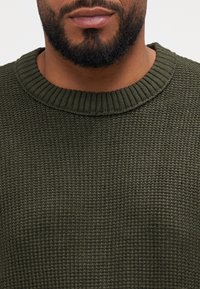 Närbild av en man med skägg som bär en texturerad, mörkgrön stickad tröja med ribbad rundad halsringning.