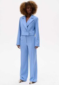 Przycięty niebieski żakiet z podwójnymi guzikami, zestawiony z szerokimi niebieskimi spodniami. Gładki materiał o dopasowanym kroju i głębokim dekolcie w kształcie V.