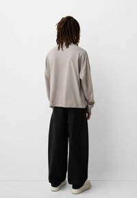 Light beige long-sleeve top with a relaxed fit, paired with wide black trousers and white sneakers, viewed from the back against a plain background.
