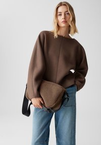 Donna bionda in un ampio maglione marrone e jeans blu tiene in mano una grande borsa a spalla marrone con texture su uno sfondo chiaro.