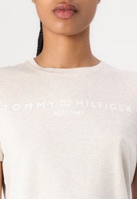 Šviesiai smėlinės spalvos medvilninis marškinėlis su trumpomis rankovėmis ir apvaliu iškirpimu, su balta "TOMMY HILFIGER EST. 1985" užrašu priekyje.
