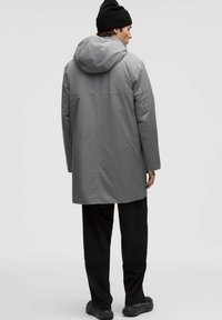 Mees hallis kapuutsiga jope, mustade pükste, musta beanie ja mustade kingadega seisab seljaga seina vastu, mis on ühtlase heleda taustaga.