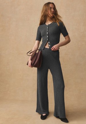 Femeie care poartă un top texturat gri închis cu mâneci scurte cu nasturi și pantaloni largi asortați, ținând o geantă maro, stând într-un decor bej.
